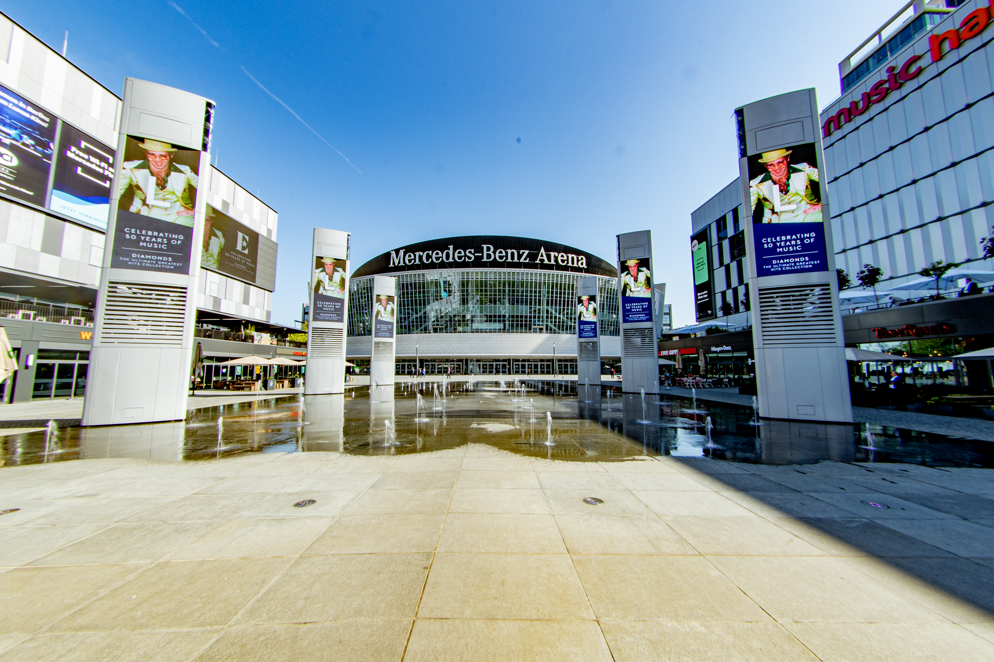 Mittig auf dem Platz vor der Mercedes-Benz-Arena stehend mit zentralem Blick auf die Glasfassade der Halle. Rechts und links stehen riesige Bildschirme mit wechselnder Werbung.